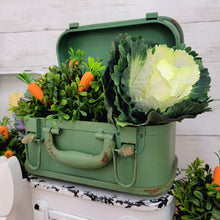 Load image into Gallery viewer, Green metal suitcase centerpiece with faux cabbage, carrots, and a boxwood orb. 
