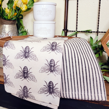 Load image into Gallery viewer, Black and off white honeybee themed cotton tea towel set with coordinating pinestripe towel displayed on a hexagon wood pedestal with ceramic sale and pepper cellars.

