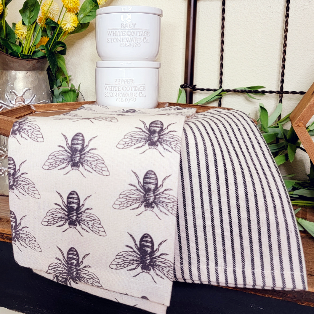 Black and off white honeybee themed cotton tea towel set with coordinating pinestripe towel displayed on a hexagon wood pedestal with ceramic sale and pepper cellars.
