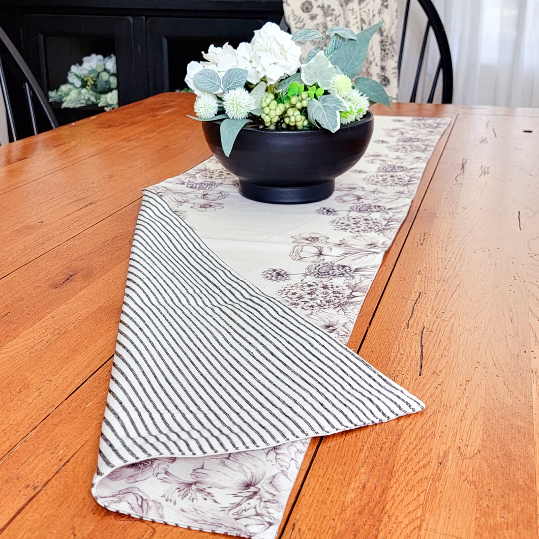 Reversible Black and White Ticking Stripe Table Runner with Sketch style botanical on the reverse. Matte black pottery bowl centerpiece with faux white hydrangea and green foliage on a farmhouse wood dining table.