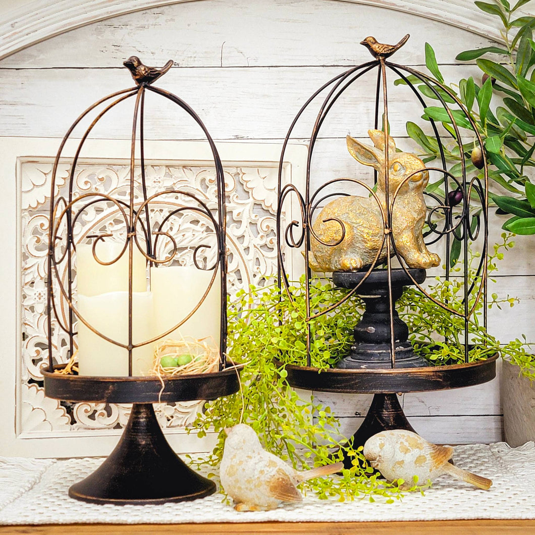 Oil rubbed brass metal birdcage cloches on stands displaying flameless pillar candles, greenery, gold easter rabbit, and weather resin bord figurines.
