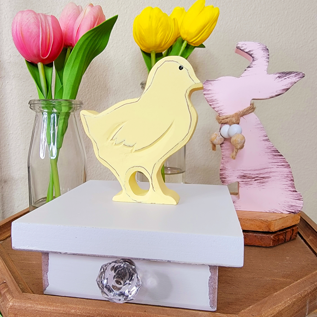 Creamy yellow wood Easter Chik tiered tray decor displayed on top of a vintage inspired white wood trinket box with a crystal knob next to a pink distressed wood bunny with beads Pink and Yellow Real Feel faux tulips in glass milk bottles