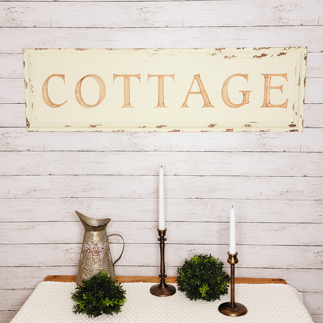 Large vintage chipped paint and distressed wood carved cottage wall decor sign hung on a shiplap wall above a sidetable displaying faux boxwood half spheres, a rustic bee embossed metal pitcher, and copper metal taper candle holders.
