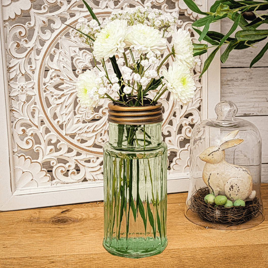 Flower frog glass vase with white floral arrangement in front of a white scroll wall decor and vintage rabbit figurine on a birds nest under a small glass cloche.