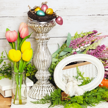Load image into Gallery viewer, Spring and Easter side table decorated with pink and yellow tulips in glass milk bottles next to a frosted silver mercury glass pillar candle holder with a birds nest on top filled with pastel colored mini glass easter ornaments. A gren fern spread out on the table with an oval white framed mirror on top with cottage bunnies peeking in.
