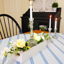 Load image into Gallery viewer, Artificial Cream Roses with Assorted White Flowers and Foliage floral picks centerpiece in a white wood trough alongside ornate glass taper candlestick holders on top of a blue and white stripe tablecloth with fringe
