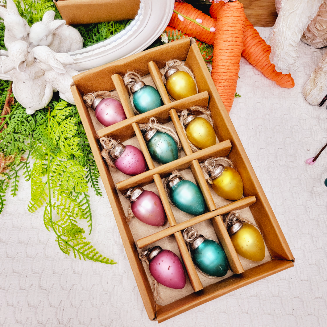 12 Miniature distrssed pastel egg shaped glass ornaments displayed in a brown box surrounded by faux ferns and Easter bunny figurines.
