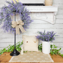 Load image into Gallery viewer, Spring Easter themed side table display featuring a lavender wreath with tan jute bow hung on a black iron wreath stand next to a linen patchwork rabbit pillow, a white ceramic pitcher used as a vase with artificial lavender picks. In the background there is sage green faux garland and a white mantel.
