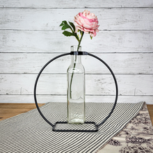 Load image into Gallery viewer, Glass bottle stem vase on a black metal stand with a single pink peony displayed on a sidetable with a black and white stripe table runner.
