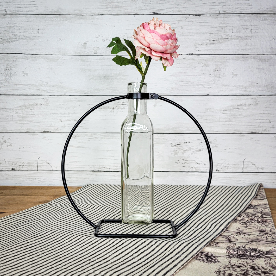 Minimalist Glass Bud Vase suspended in a circular black metal stand with an artificial pink peony setting on top of a black and white ticking stripe table runner
